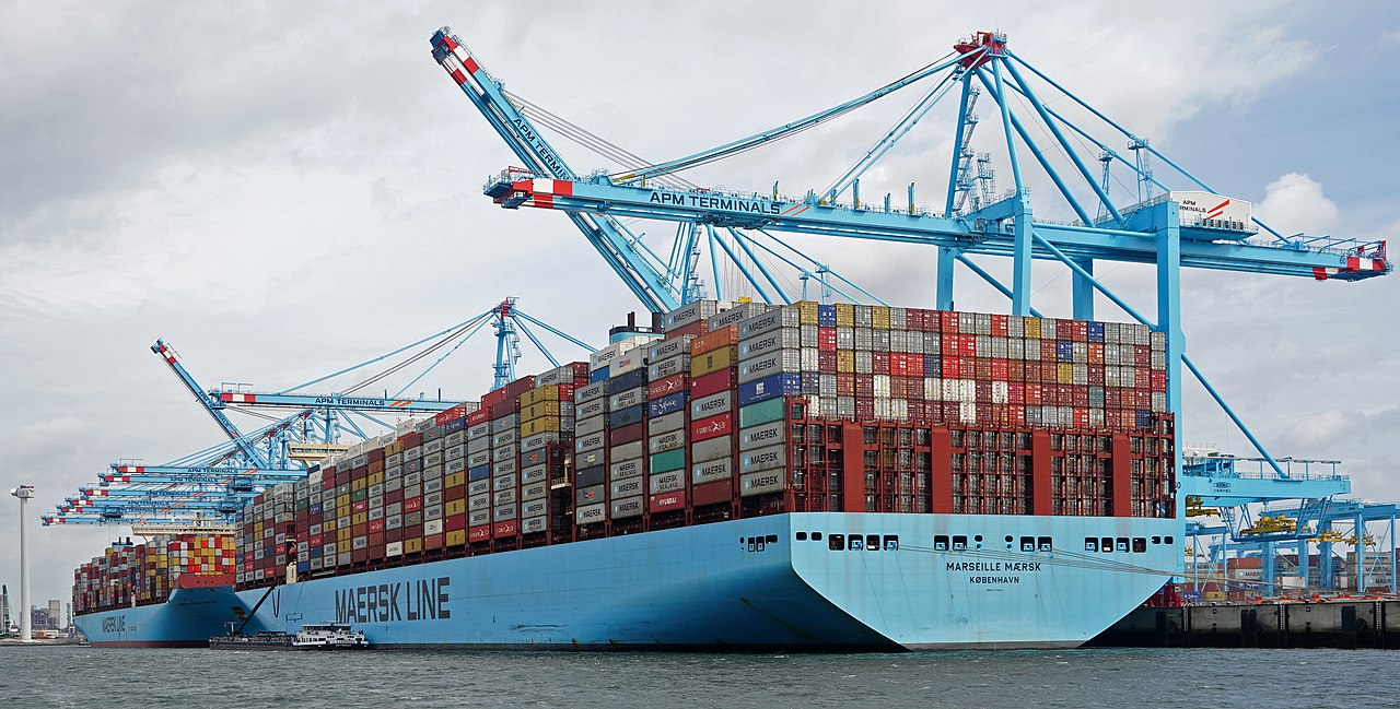 Ett stort containerfartyg vid kaj under hamnkranar, lastat med containrar i flera våningar.