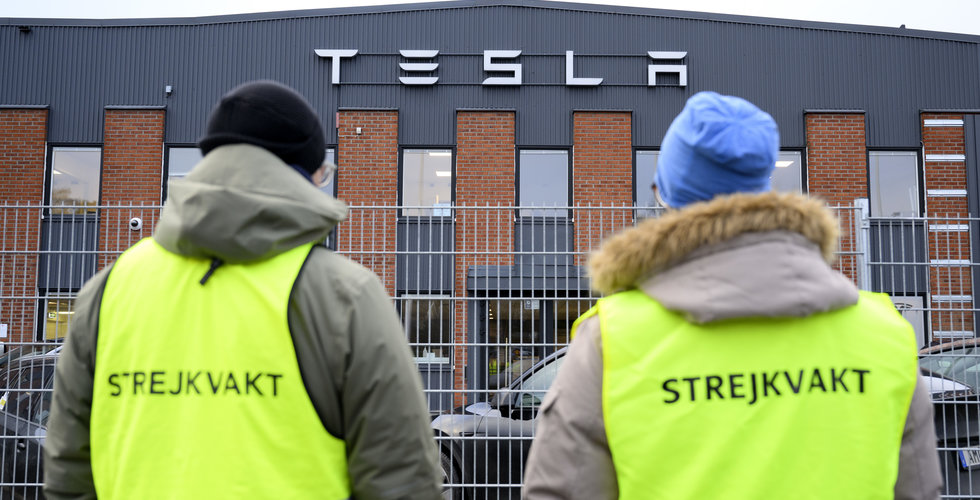Två strejkvakter i gula västar står utanför en byggnad med Teslas namn på fasaden.